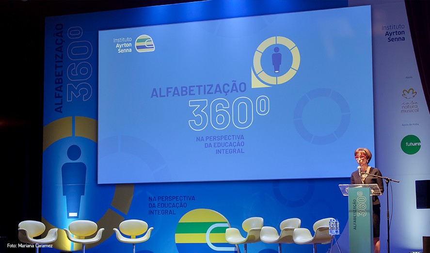 Uma mulher em um pódio em frente a uma tela durante um evento focado na ‘alfabetização 360º’.