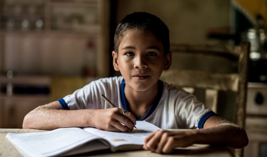 Um menino está sentado à mesa e escrevendo em um caderno como parte da nova fase da Volta ao Novo com o Consed, que tem como foco mapear boas práticas educacionais.