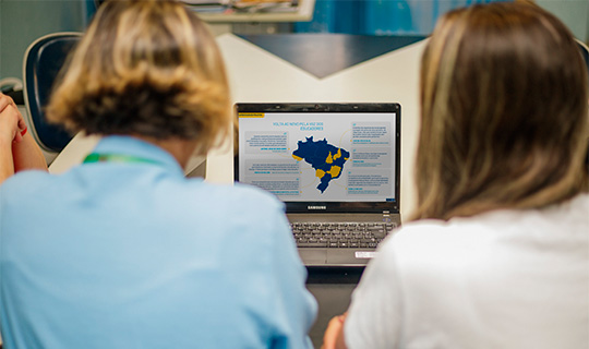 Duas mulheres sentadas à mesa olhando para um laptop com um mapa do Brasil, representando "Quem Somos".