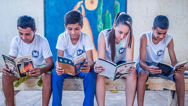 Na Sala De Aula, um grupo de crianças sentadas num banco lendo livros.