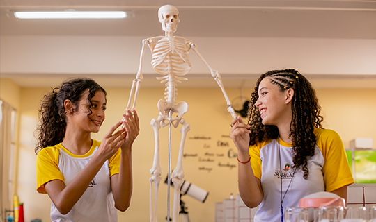 Duas meninas ao lado de um esqueleto em uma sala de aula do Instituto Ayrton Senna.