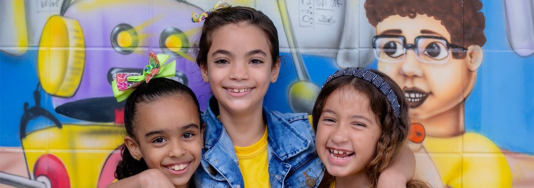 Três meninas posando para foto em frente ao mural de Pesquisas Sobre Socioemocionais.