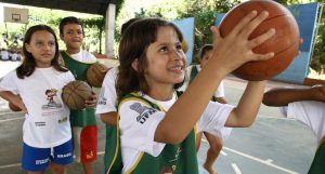 Um grupo de crianças pratica Educação pelo Esporte, jogando basquete em uma academia.