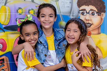 Três meninas posando para foto em frente a um mural durante Pesquisas Sobre Socioemocionais.