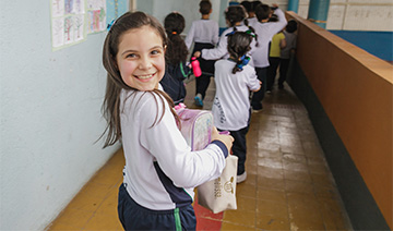 Uma menina caminha por um corredor segurando um livro, rodeada de componentes educativos.