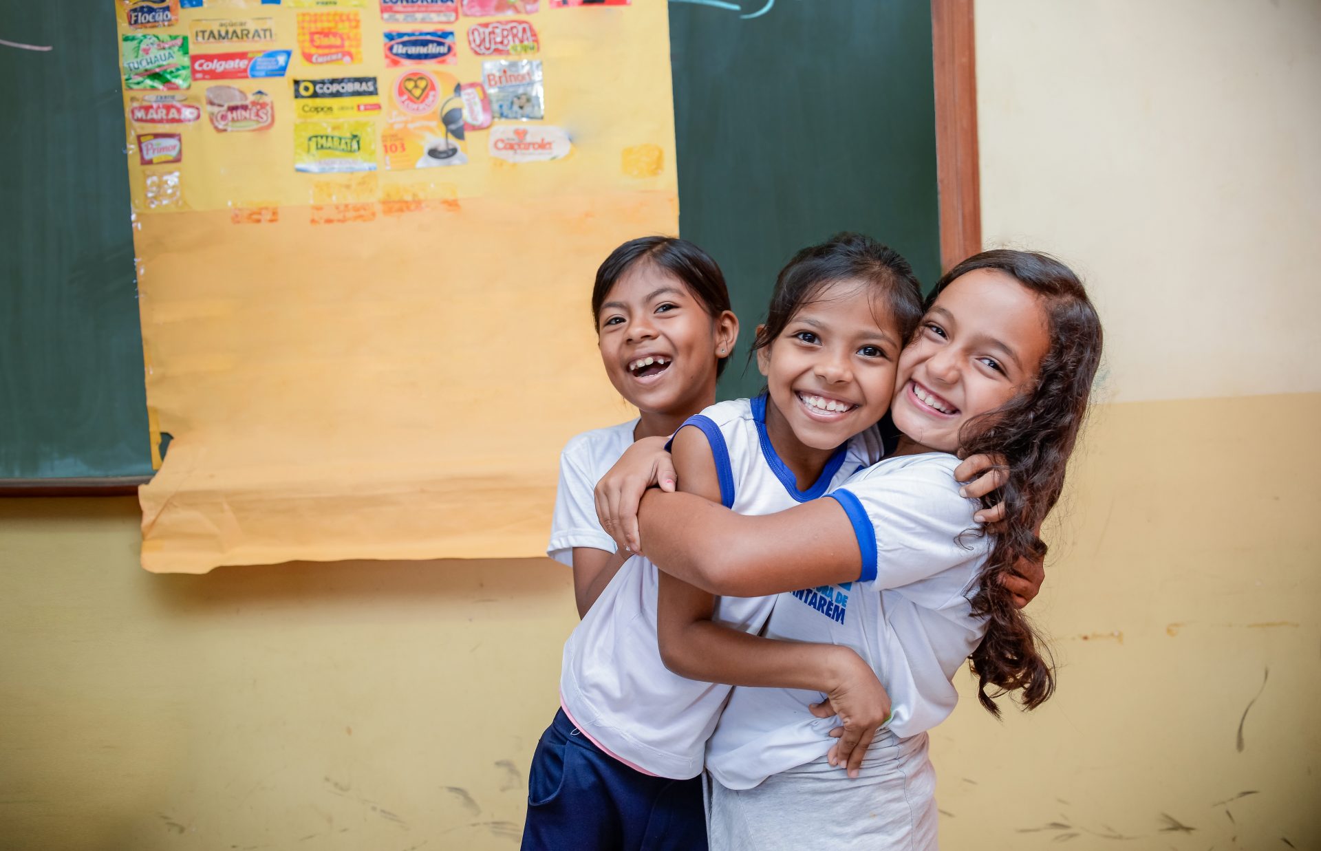 Três meninas se abraçando em frente a um quadro negro. Ajude o Instituto Ayrton Senna e apoie a educação fazendo uma doação.