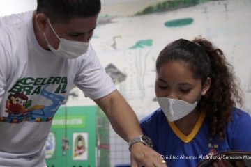 Com o apoio do Instituto Ayrton Senna, um homem usando máscara ajuda uma menina em uma sala de aula enquanto a rede municipal de Manaus reduz o índice de distorção idade-série de