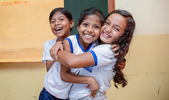Três meninas se abraçando em frente a uma sala de aula, recebendo doações internacionais do Instituto Ayrton Senna.