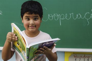 Um menino segurando um livro em frente a um quadro negro recebe um Cartão Itaú.