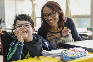 A woman in glasses smiles at a boy in a wheelchair in a classroom, providing support for education.