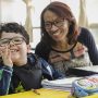 Uma mulher de óculos sorri para um menino cadeirante em uma sala de aula, promovendo apoio à educação por meio do Instituto Ayrton Senna.