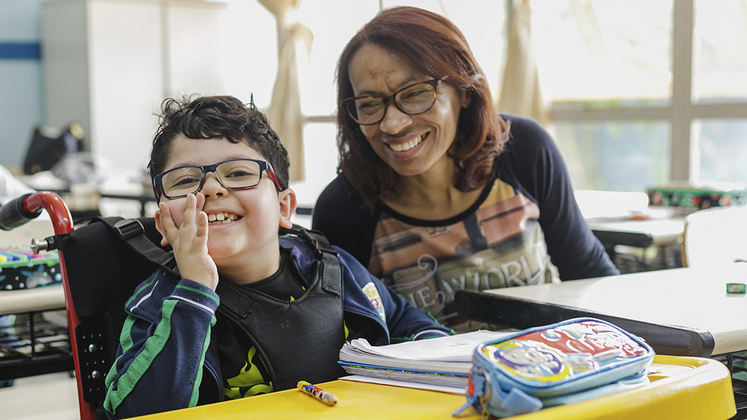 A woman in glasses smiles at a boy in a wheelchair in a classroom, providing support for education.