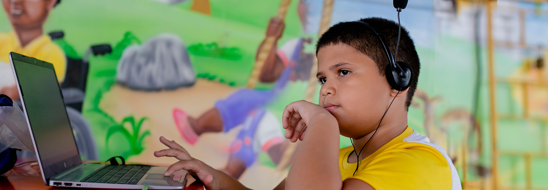 Um menino com fones de ouvido e um laptop em frente a um mural, interagindo com materiais educativos.