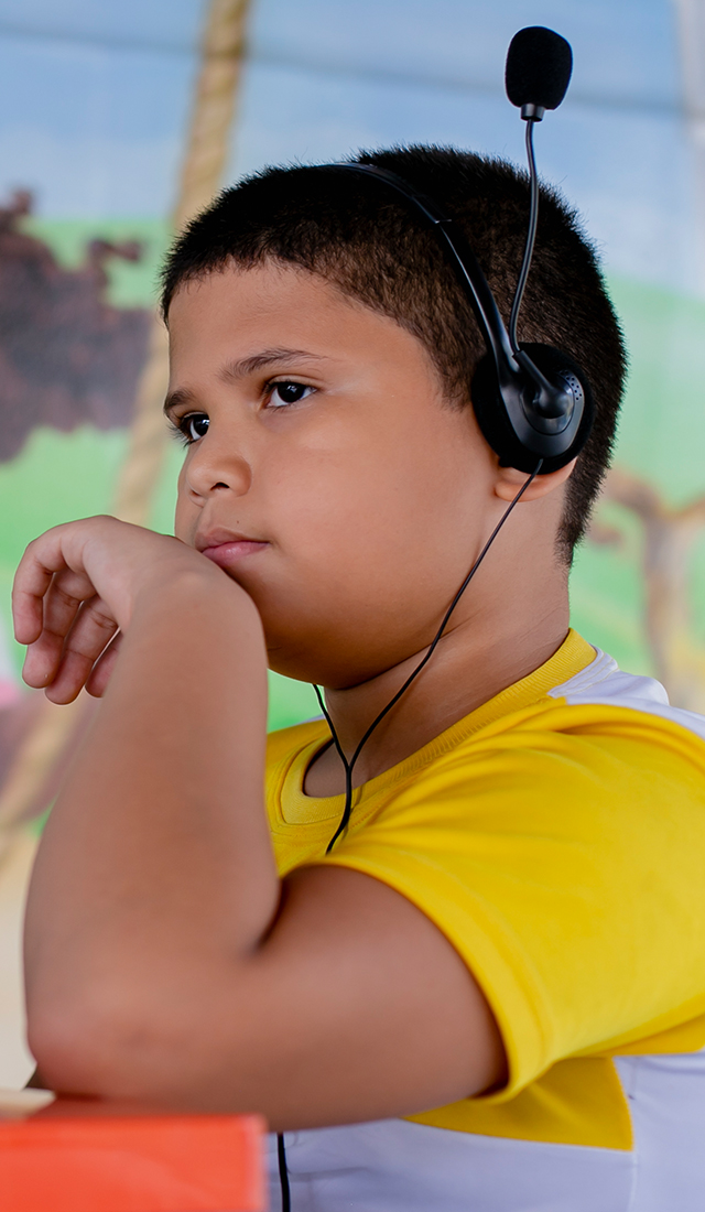 Um menino com fones de ouvido e um laptop em frente a um mural, interagindo com materiais educativos.