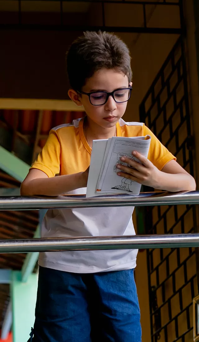 O Que Fazemos - Um menino está lendo um livro em pé sobre uma grade.