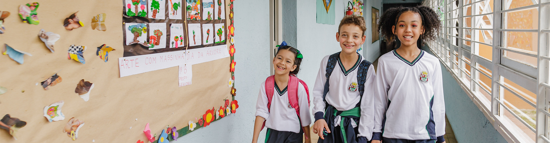 Alunos sorrindo chegando na escola andando pelo corredor com mochilas e materiais