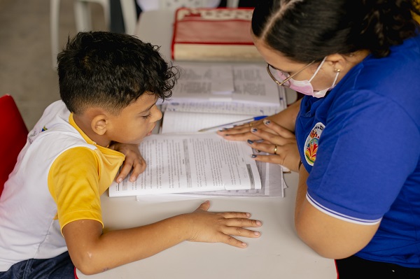 Professora ensinando aluno em uma escola. Estão sentados em um carteira lendo juntos um livro adequado a BNCC seus impactos e diretrizes.