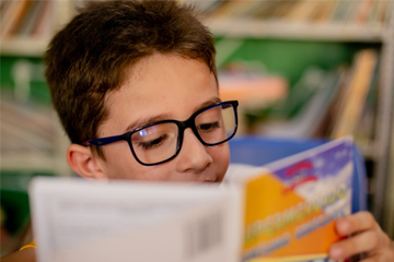 Um menino de óculos está lendo um livro em uma biblioteca, com foco na alfabetização.