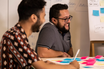 Um grupo de pessoas sentadas ao redor de um quadro branco com post-its, discutindo e traçando estratégias para melhorias sociais.