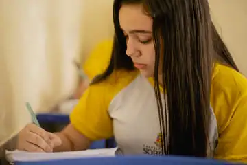 Uma garota de camisa amarela escrevendo em um caderno, preparando-se para os desafios e oportunidades do PISA.