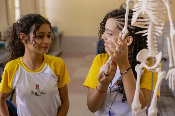 Duas meninas ao lado de um esqueleto em uma sala de aula, servindo de modelo para profissões.