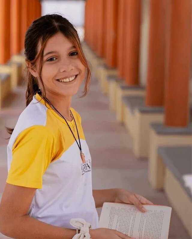 Uma menina vestindo uma camiseta amarela e branca do Instituto Ayrton Senna e segurando um livro.