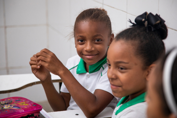 Duas meninas com uniforme escolar sentadas em uma mesa, uma delas sorrindo para a câmera, em sala de aula durante o censo escolar.
