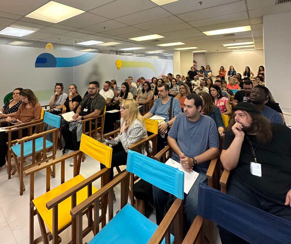 Um grupo diversificado de adultos sentados em uma sala com cadeiras assistindo atentamente a uma apresentação sobre competências socioemocionais.