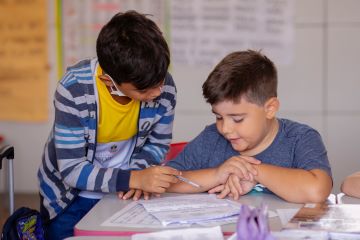 Dois meninos sentados em uma mesa de sala de aula, trabalhando juntos em uma planilha, com cartazes socioemocionais e educativos ao fundo.