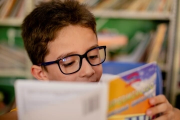 Uma criança de óculos engajada intensamente na alfabetização lendo um livro em um ambiente de biblioteca.