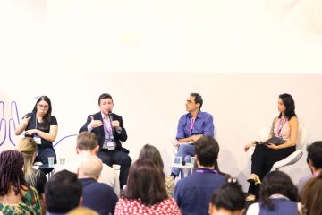 Na Bett Brasil, quatro painelistas discutiram em uma conferência diante de uma plateia, com um homem falando e outros três ouvindo atentamente..