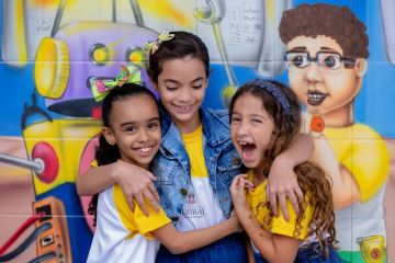 Três crianças sorrindo e se abraçando diante de um mural colorido com elementos lúdicos e artísticos em Sobral, palco do estudo da OCDE.