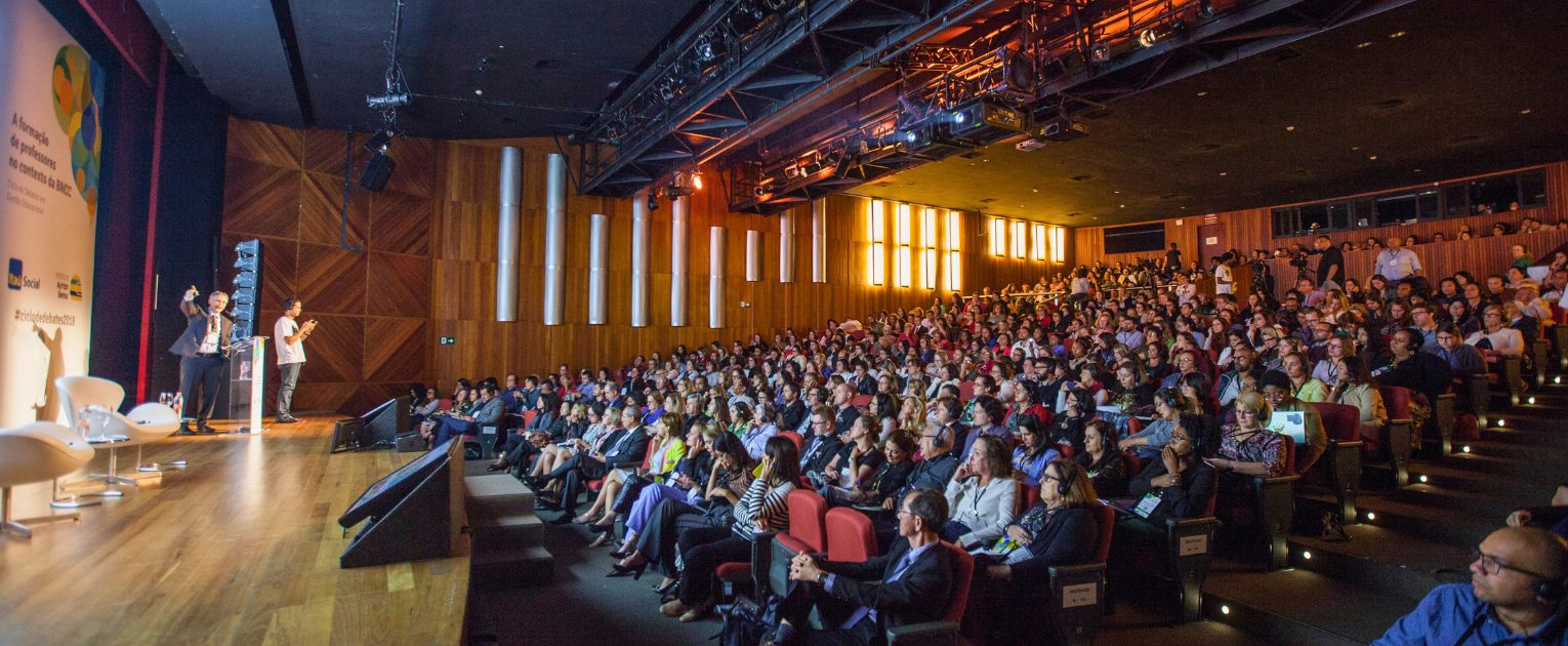 Um grande público sentado em um auditório moderno ouve um palestrante em um pódio durante uma conferência.