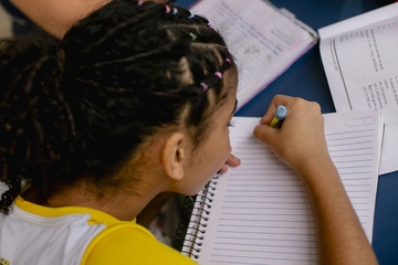 A student with braided hair writing in a notebook during class, with textbooks and papers scattered around on the desk.