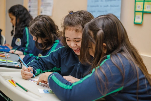 Quatro meninas com uniformes escolares sentadas em uma mesa, rindo e trabalhando juntas em uma tarefa em uma sala de aula, otimizada para SEO.