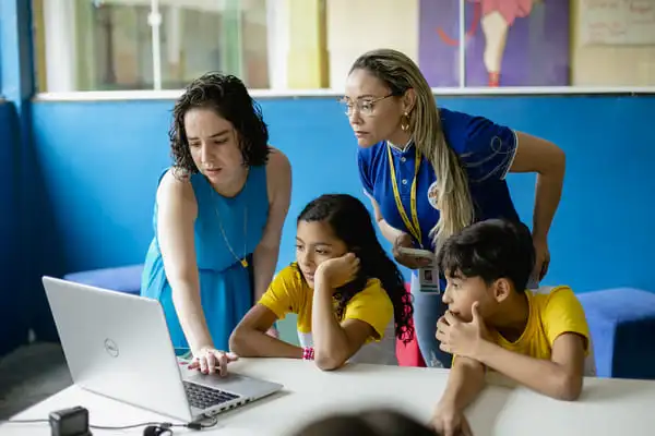 Duas mulheres e duas crianças olham para a tela de um laptop em uma sala de aula, participando de uma atividade educacional.