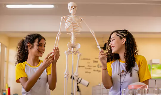 Dois estudantes de camisa amarela examinando um modelo de esqueleto humano em uma sala de aula do Instituto Ayrton Senna.