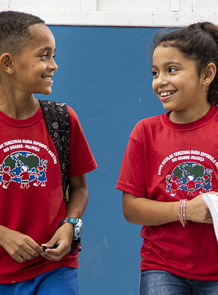Duas crianças de camisa vermelha sorrindo e conversando, com uma parede azul ao fundo. Uma criança usa uma mochila.