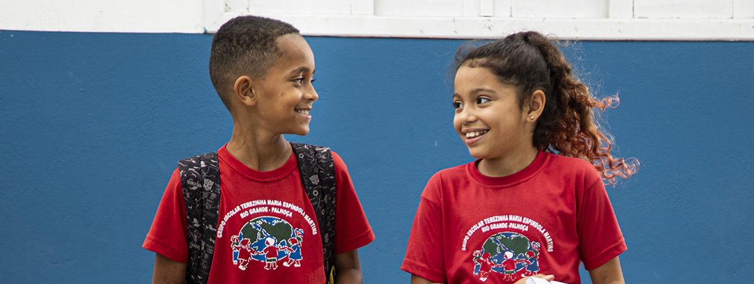 Duas crianças, um menino e uma menina, sorrindo um para o outro, usando mochilas e camisetas escolares vermelhas fornecidas por doações por empresas, em frente a uma parede azul.