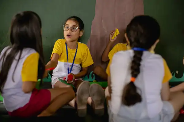 Uma jovem de óculos e camisa amarela fala animadamente sobre educação socioemocional para três colegas durante uma atividade em grupo em uma sala de aula.