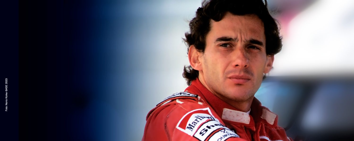 A close-up of a male race car driver in a red and white uniform, looking pensively to the side under bright lighting.