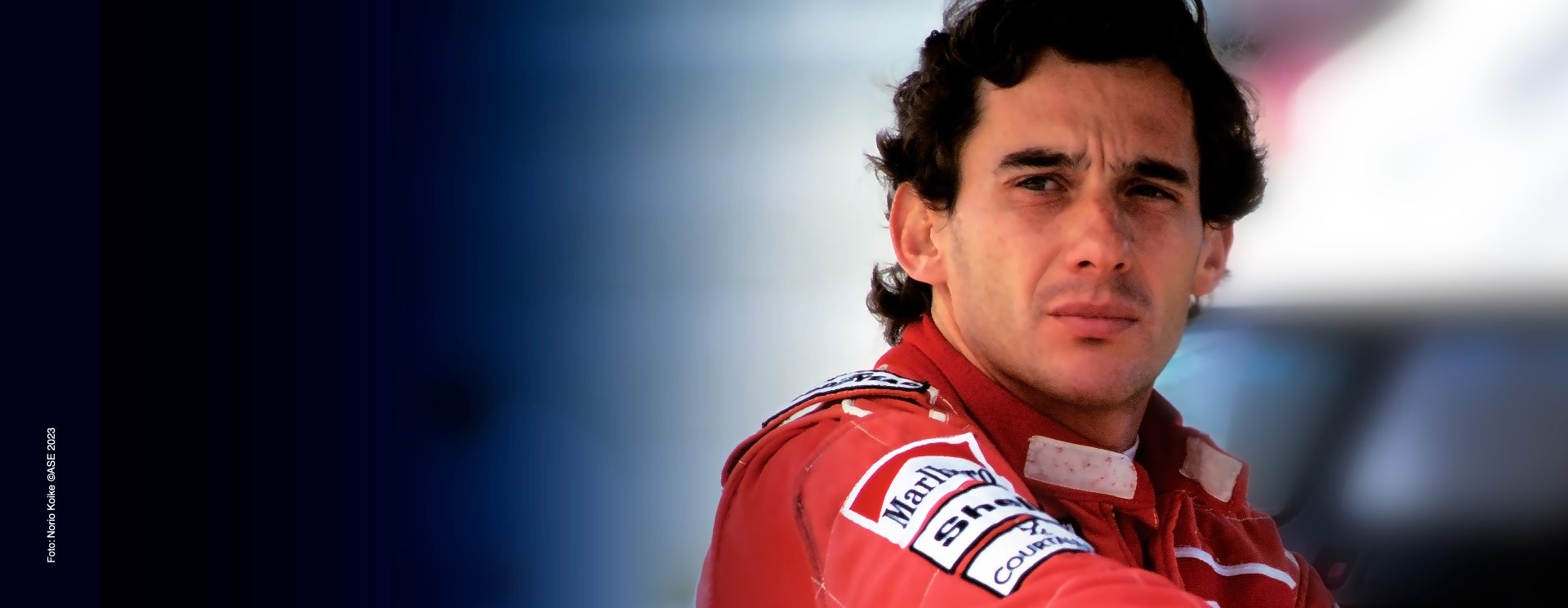 Retrato do piloto de corrida Ayrton Senna em um macacão vermelho, parecendo pensativo com um fundo de pista desfocado.