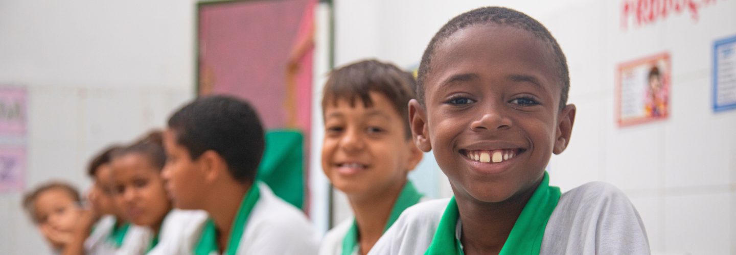 Alunos em uniforme escolar sorrindo em uma sala de aula, com foco em um menino alegre em primeiro plano segurando um Cartão Instituto Ayrton Senna Itaú.