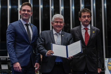 Três homens de terno, um deles com certificado, juntos e sorrindo, em um ambiente formal que lembra uma cerimônia de premiação de Ayrton Senna.