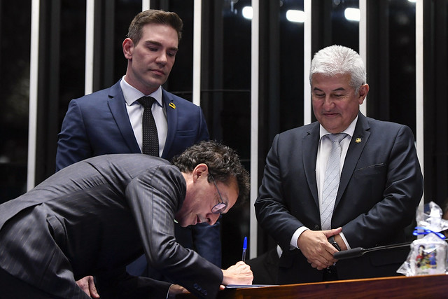 Um homem assina um documento em uma mesa de reunião, observado por dois homens em pé, um deles segurando uma pasta, em homenagem a Ayrton Senna.