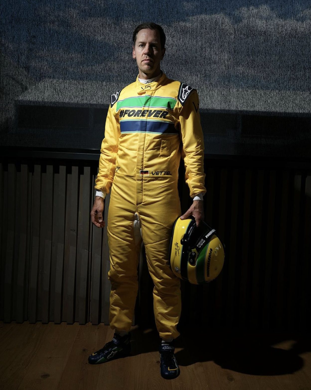 Uma pessoa com um traje de corrida amarelo segura um capacete, evocativo do equipamento icônico de Ayrton Senna. O traje tem “#ForEver” estampado no peito. O fundo é escuro e interno.