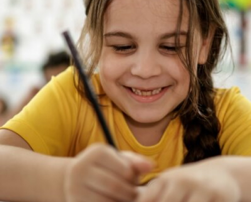 Uma jovem de trança sorri enquanto escreve a lápis no papel, vestindo uma camisa amarela, sonhando onde estará daqui a 30 anos.