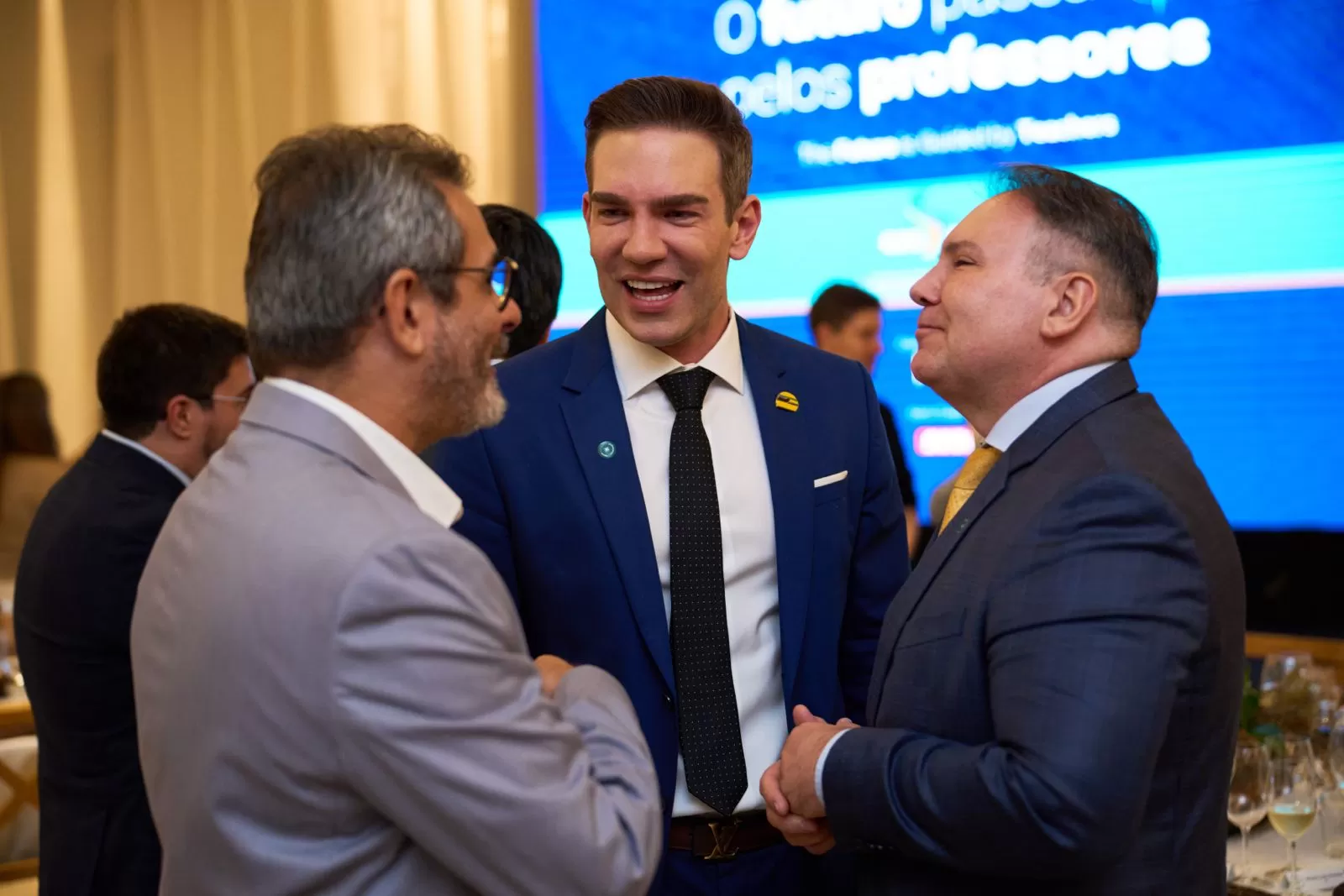 Três homens de terno conversam em um evento formal, com uma tela azul de apresentação durante o G20.