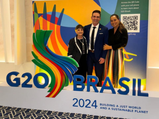 Três pessoas sorriem em frente a uma placa do G20 Brasil 2024 com um logotipo colorido. A placa, quase como um rascunho automático, diz “Construindo um Mundo Justo e um Planeta Sustentável”.