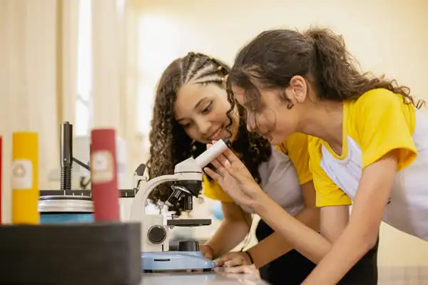 Duas meninas em uma sala de aula examinando uma amostra ao microscópio, sorrindo e engajadas em uma observação científica.